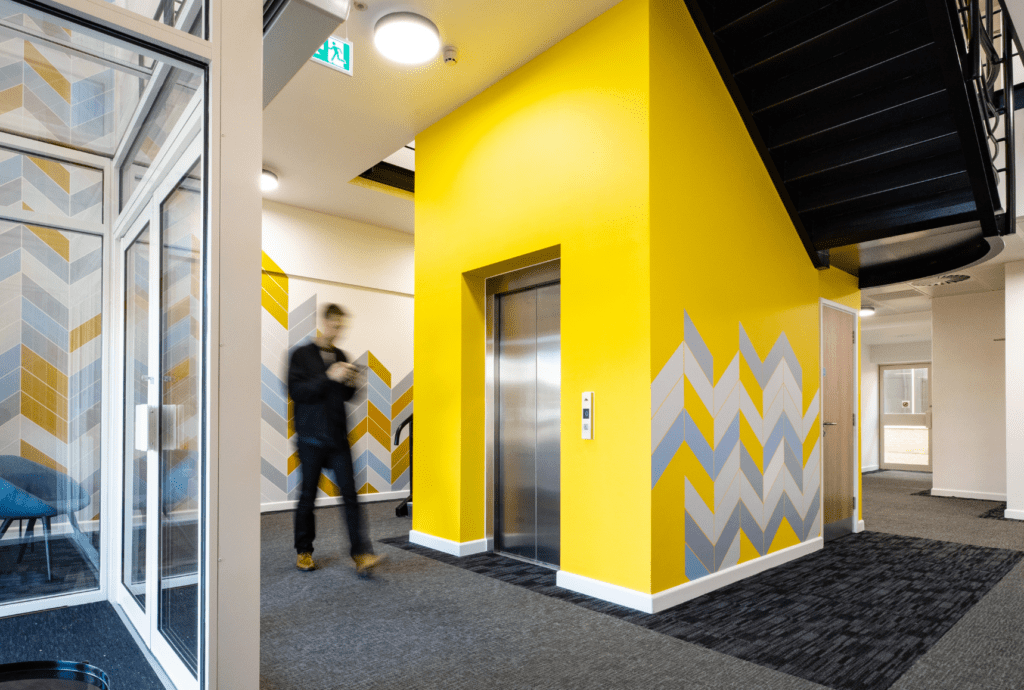 Man walking past a lift in a CAT A fit-out commercial office space at Pinesgate in Bath. The walls are yellow and there is a grey carpet.