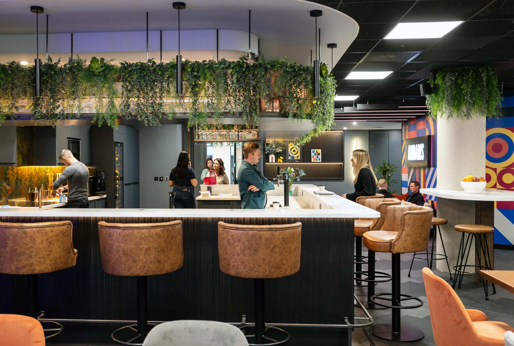 Modern office bar and breakout space with leather bar stools and colourful wall murals, with people chatting at the bar