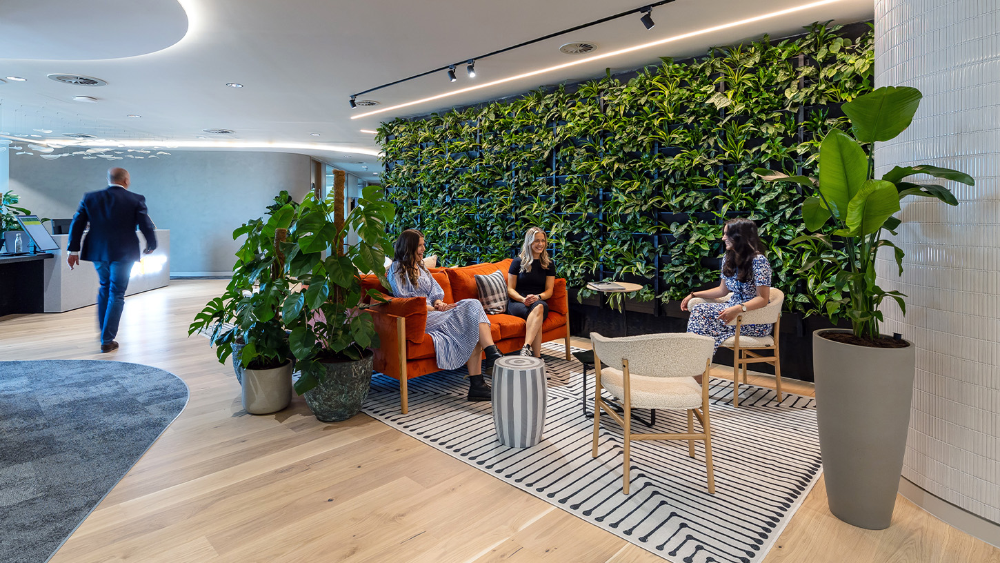Office welcome area with cosy orange sofa, cream sherpa armchairs and living plant wall with three women chatting