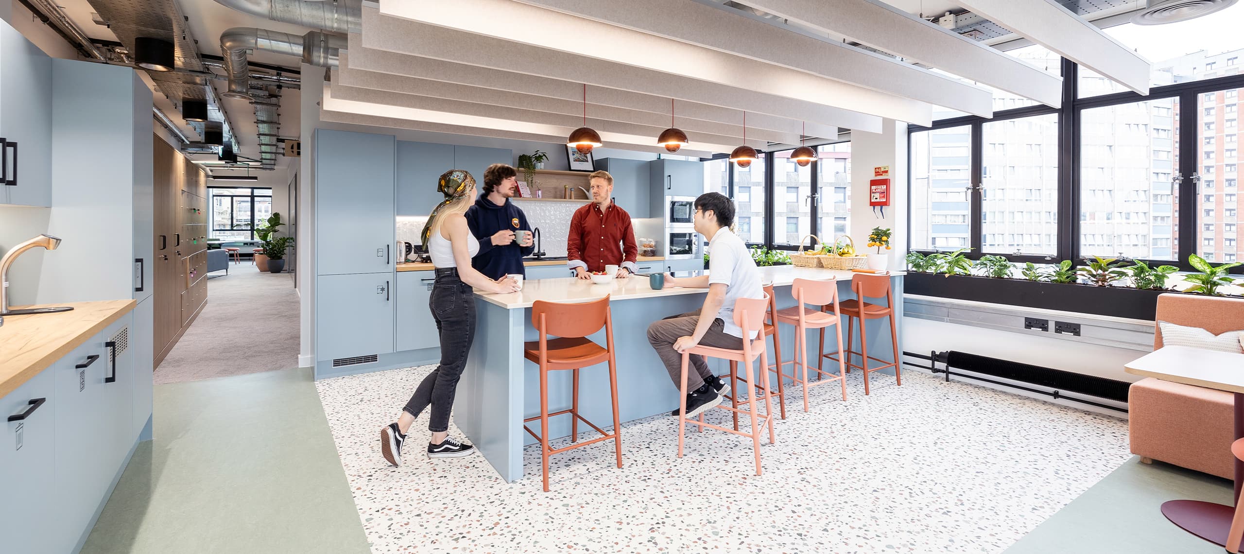 A group of people stand at a light blue kitchen counter chatting, with pink bar stools