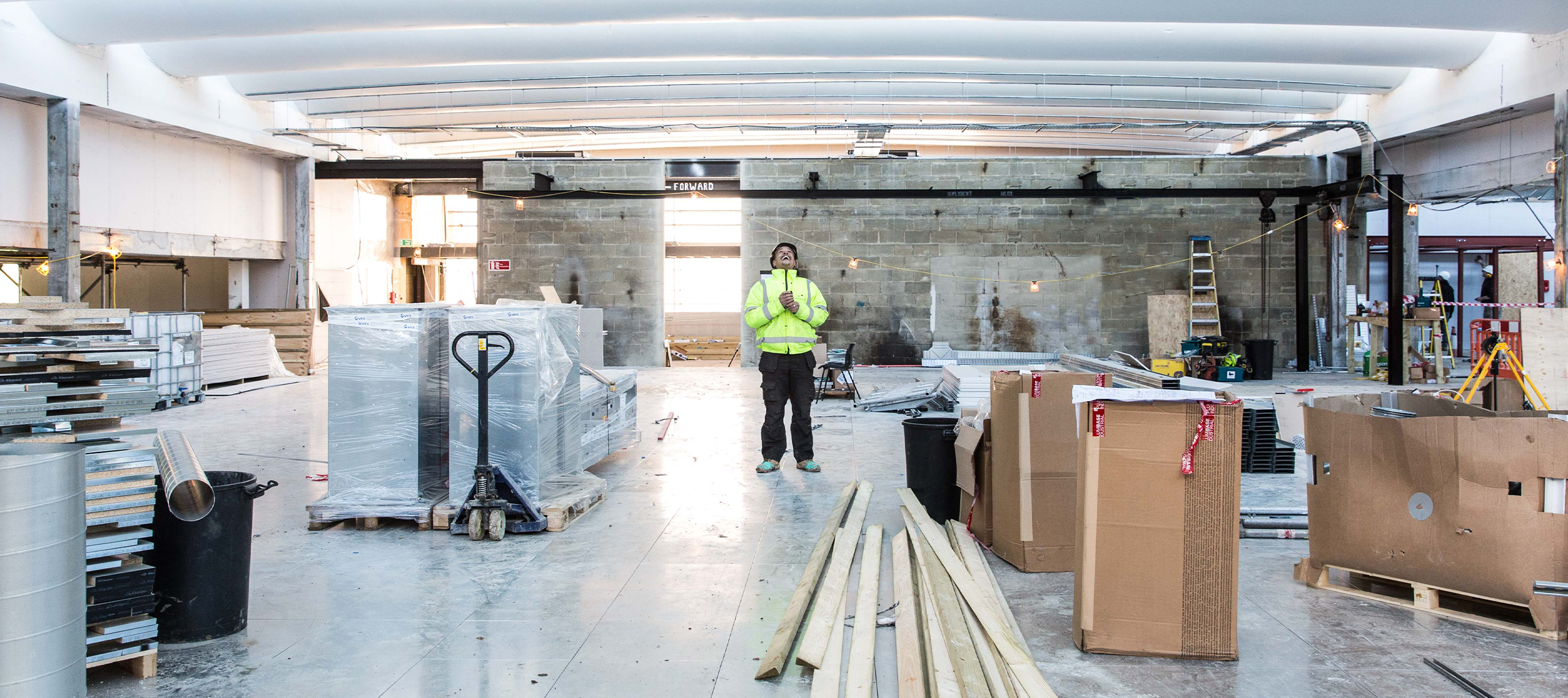A site manager in hivis and hard hat smiles in the middle of a building site