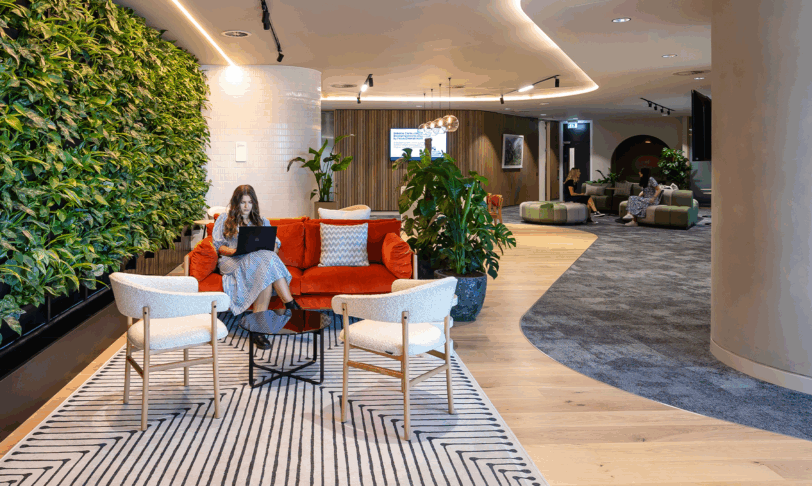 A modern office with woman working on a laptop sat on a cosy bright orange sofa next to a living plant wall