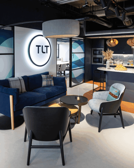 A comfy seating area in an office with navy blue sofa and the TLT logo lit up on the wall, office kitchen in the background