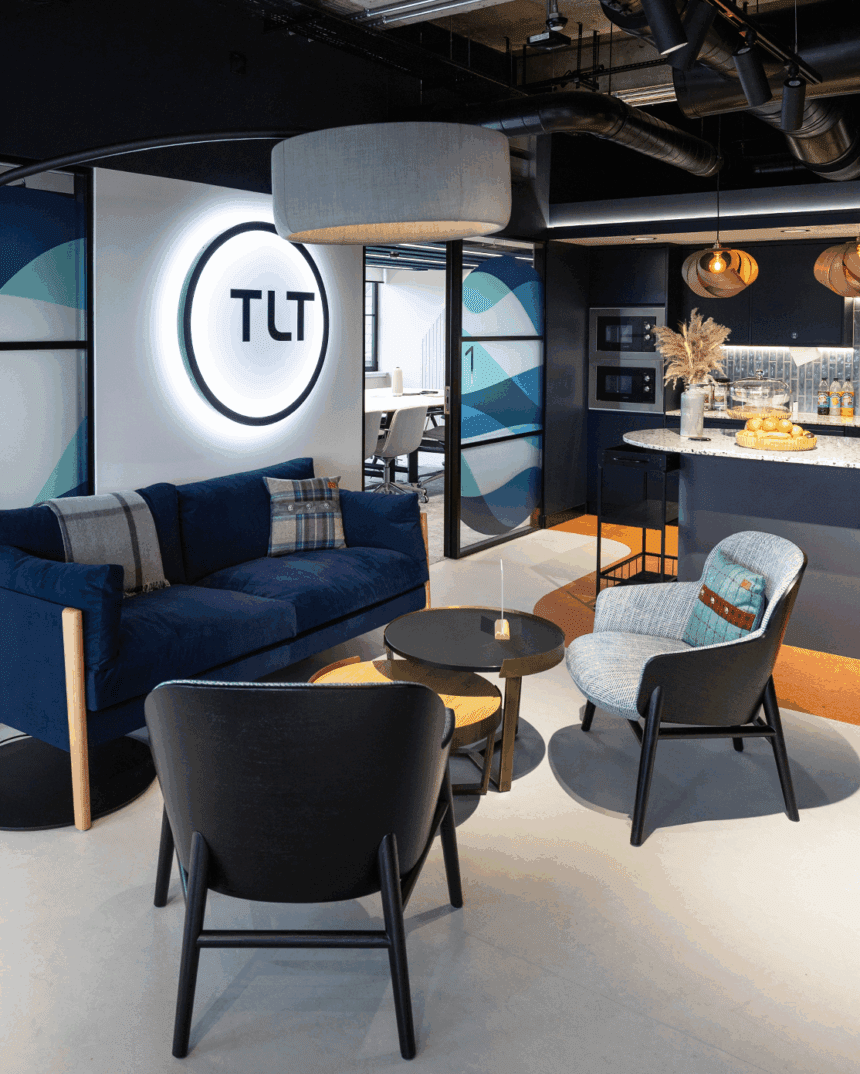 A comfy seating area in an office with navy blue sofa and the TLT logo lit up on the wall, office kitchen in the background