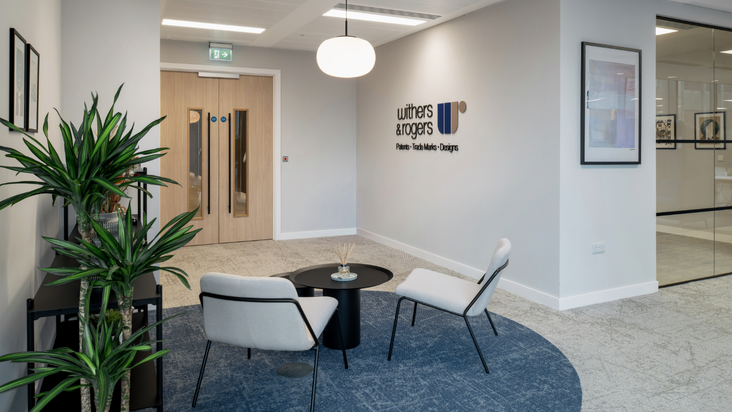 A welcoming reception area, with soft beige seating and blue curved carpets, with shelving to display patented products and green foliage.
