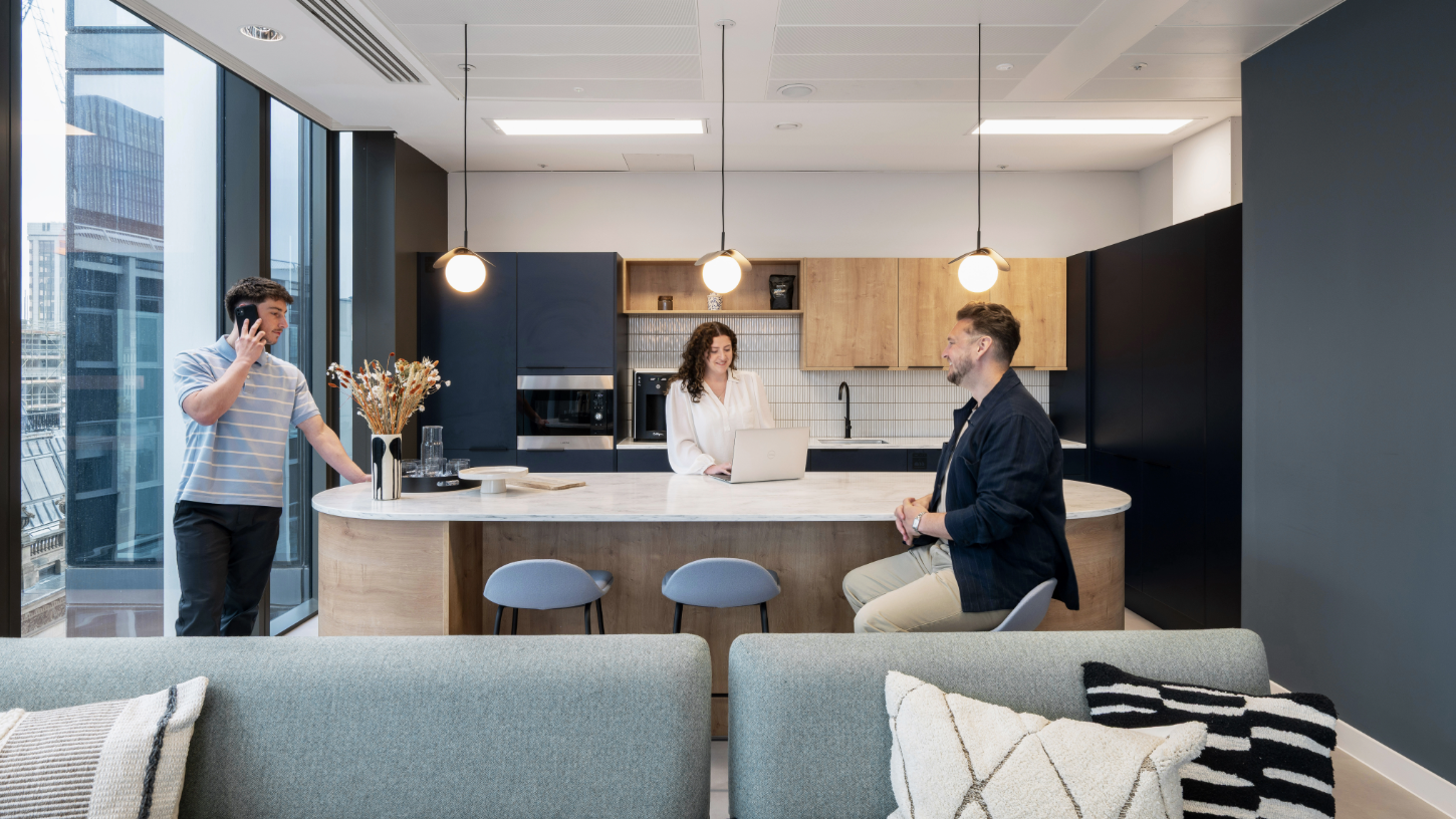 A communal kitchen area with three people conversing over the breakfast bar, framed by a duck egg blue sofa and soft bulb lighting.