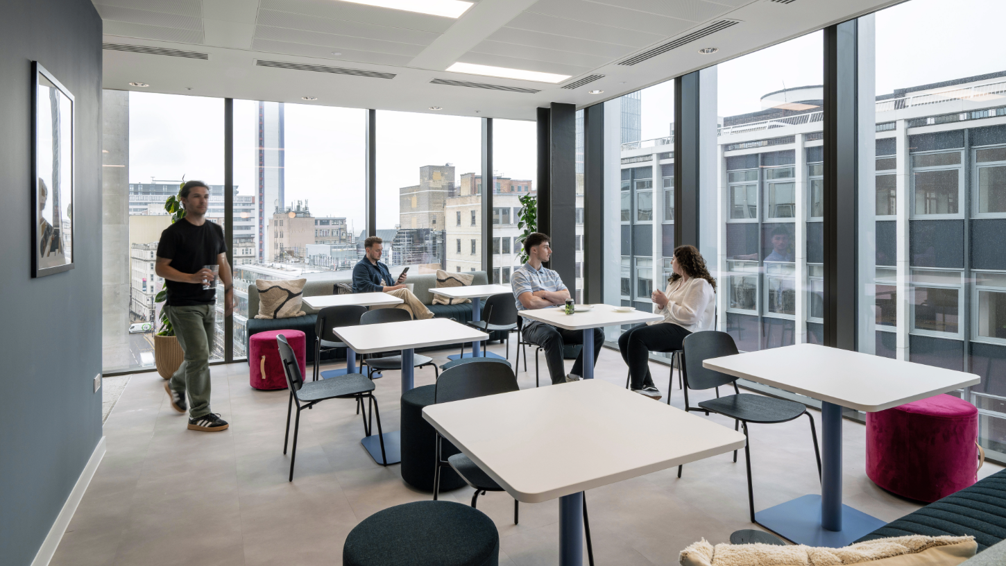 An open plan breakout space with lunch tables, and floor to ceiling windows with views over the city.